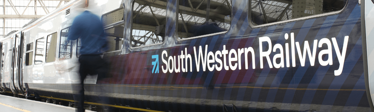 A South Western Railway class 159 diesel "West of England" train. A man in dark trousers and a blue jumper is walking in front of it.