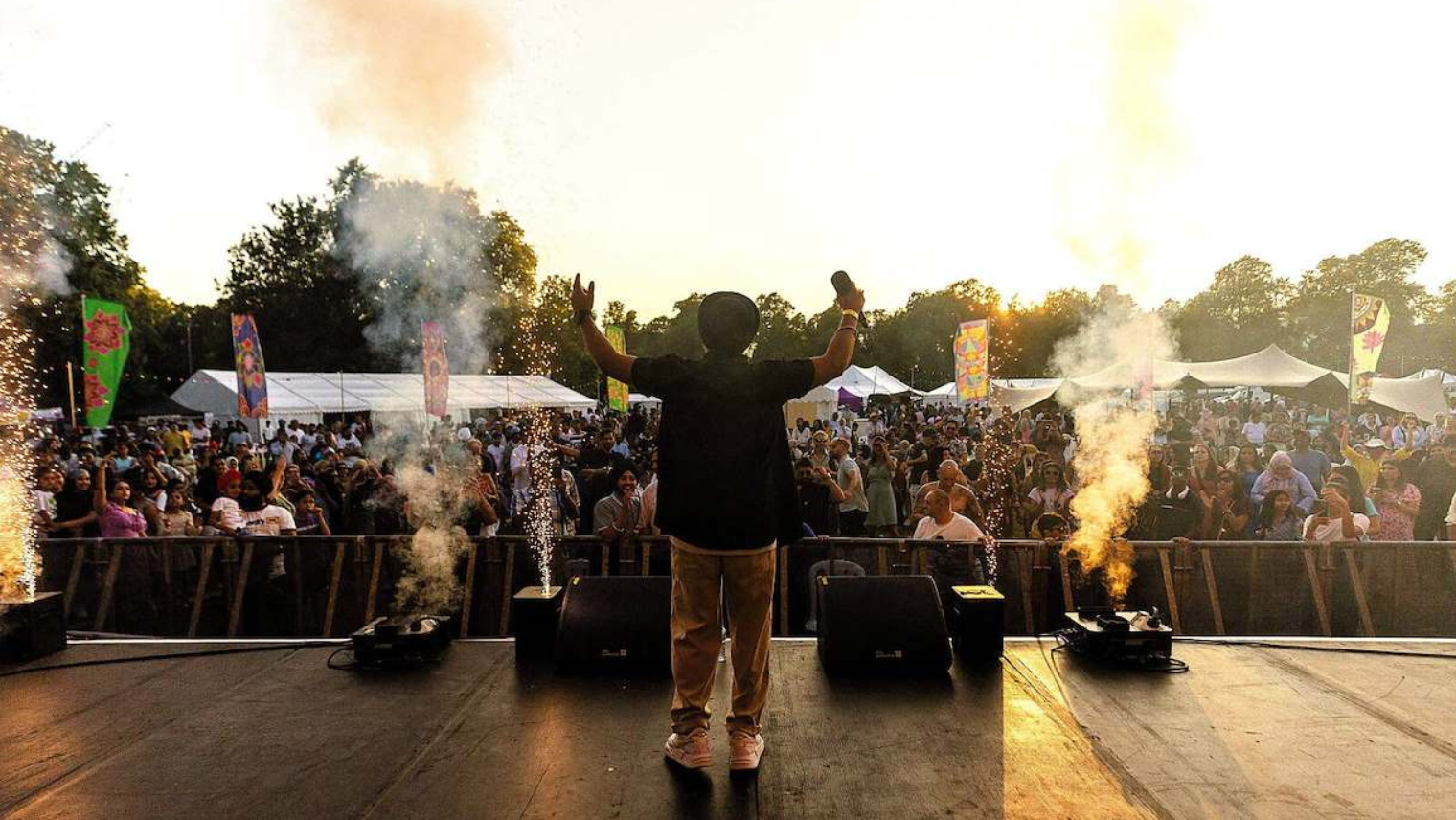Singer on stage facing a cheering crowd with fireworks and smoke effects at sunset.