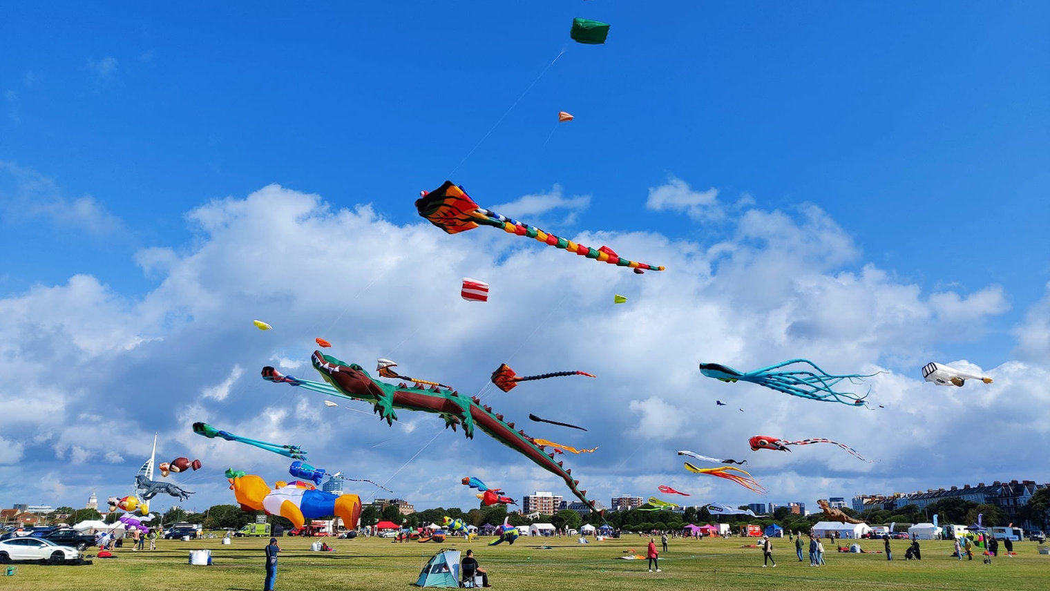 Colourful kites flying over Southsea Common, shaped like dragons, octopuses, and animals against a bright blue sky.