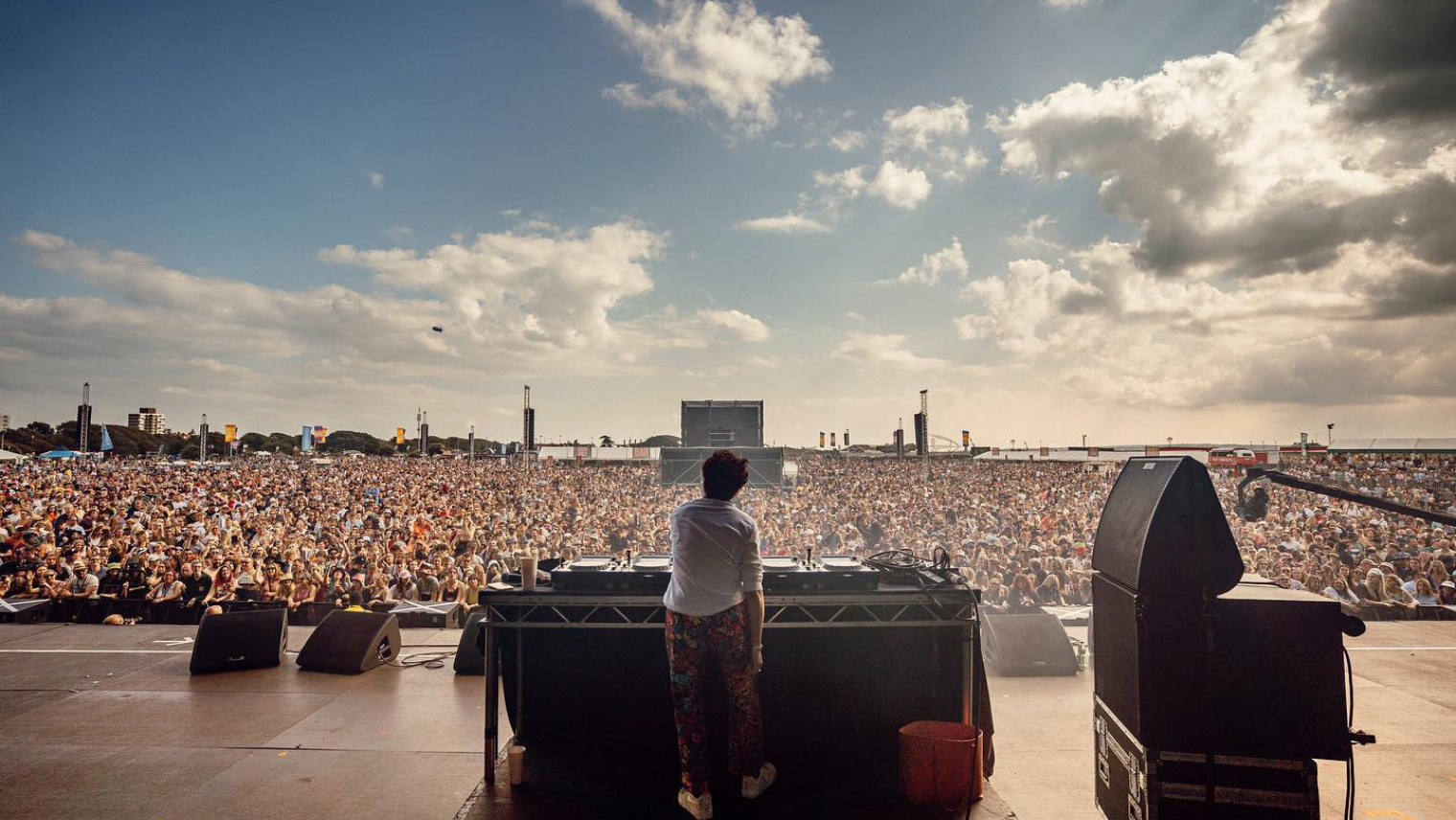 DJ performing to a huge crowd at a seaside festival, with sunlight breaking through clouds over the stage.