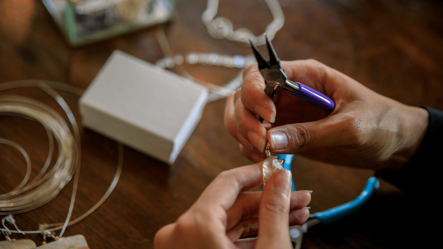 Hands use purple-handled pliers to attach a clasp to a piece of jewelry on a wooden table scattered with wire and tools.