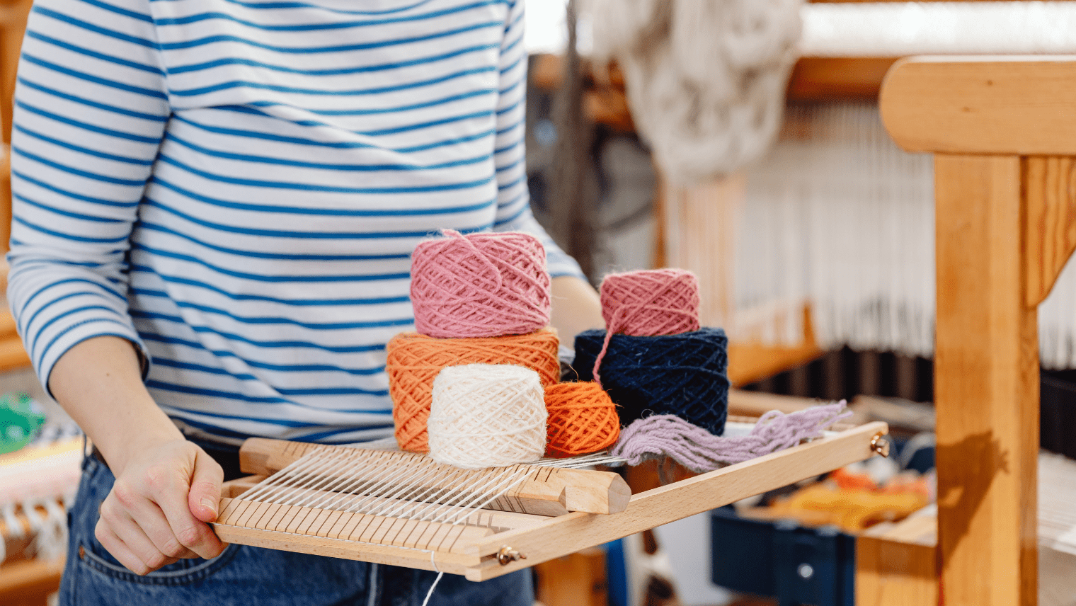 A person in a blue-and-white striped shirt holds a wooden loom with colorful yarn balls—pink, orange, white, navy, and lavender—in a textile workshop.