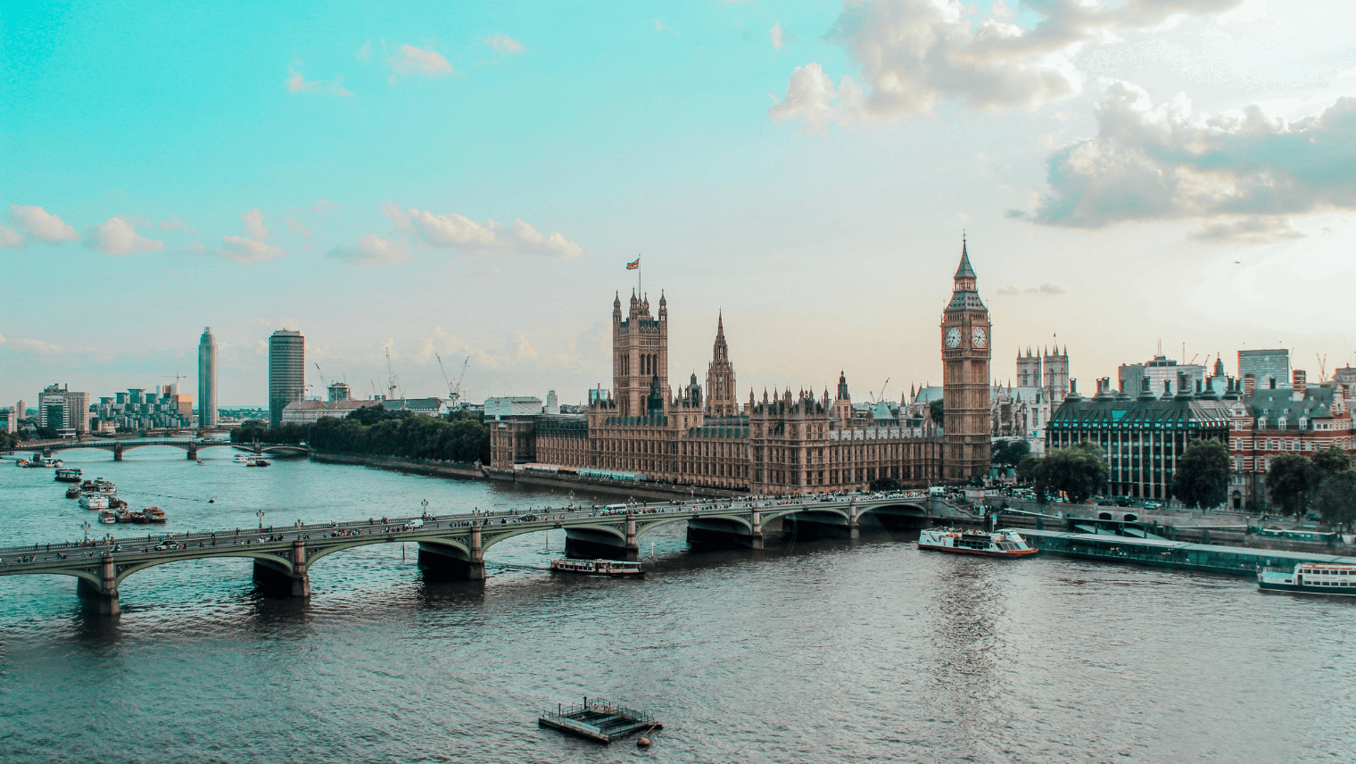 The Palace of Westminster and Big Ben overlook the River Thames under a bright sky, with boats and bridges in view.