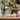 Two people wearing light green aprons arrange flowers; one cuts pink carnations with purple scissors while the other holds white lilies and carnations. Pink roses lie on the table in front of them.