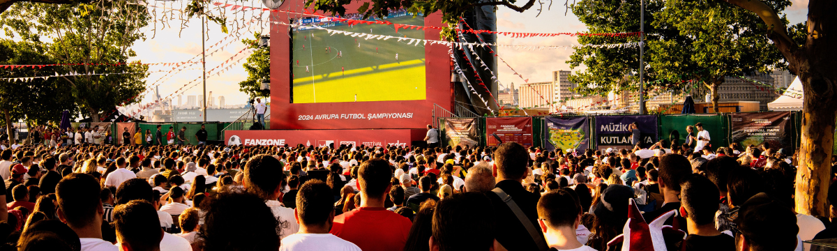 Crowd watching a football match on a big outdoor screen at a fan zone.