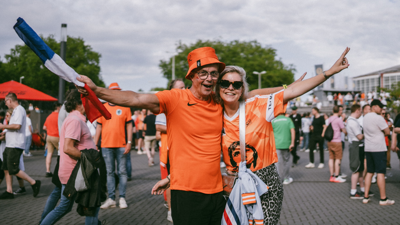 Two fans in orange outfits celebrating outdoors with a Netherlands flag among a crowd.